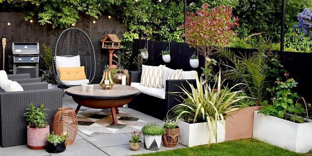 Inviting backyard patio of an Accessory Dwelling Unit with contemporary woven furniture, vibrant potted plants, a wooden round table, and a barbecue setup, surrounded by lush greenery and privacy fencing.