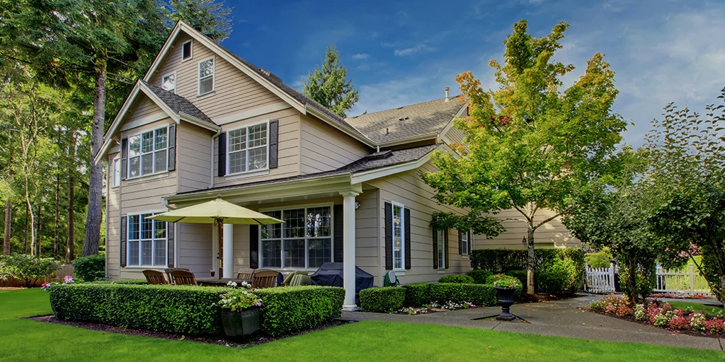 raditional two-story family home with beige siding and white trim, featuring a well-manicured lawn, colorful flower beds, and a cozy front porch with seating and a sun umbrella, nestled in a peaceful, tree-lined neighborhood.